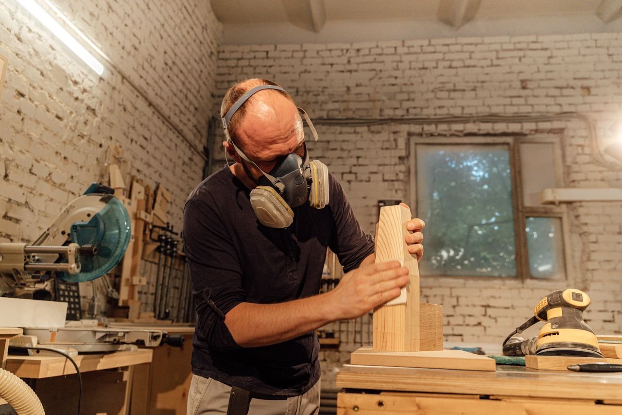 A skilled carpenter wearing a respirator mask sands a wooden piece in his workshop.