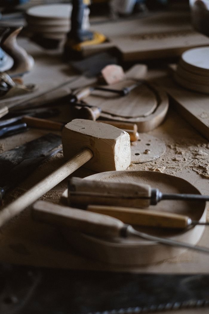 Set of chisels and wooden hammer for woodwork placed on messy table with various instruments and stack of lumber planks in joinery on blurred background
