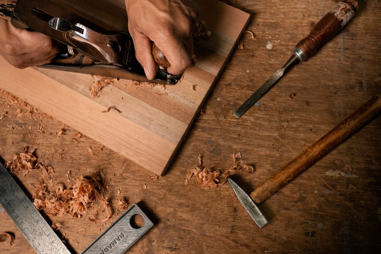 Close-up of hands using a planer on wood with tools and shavings around.