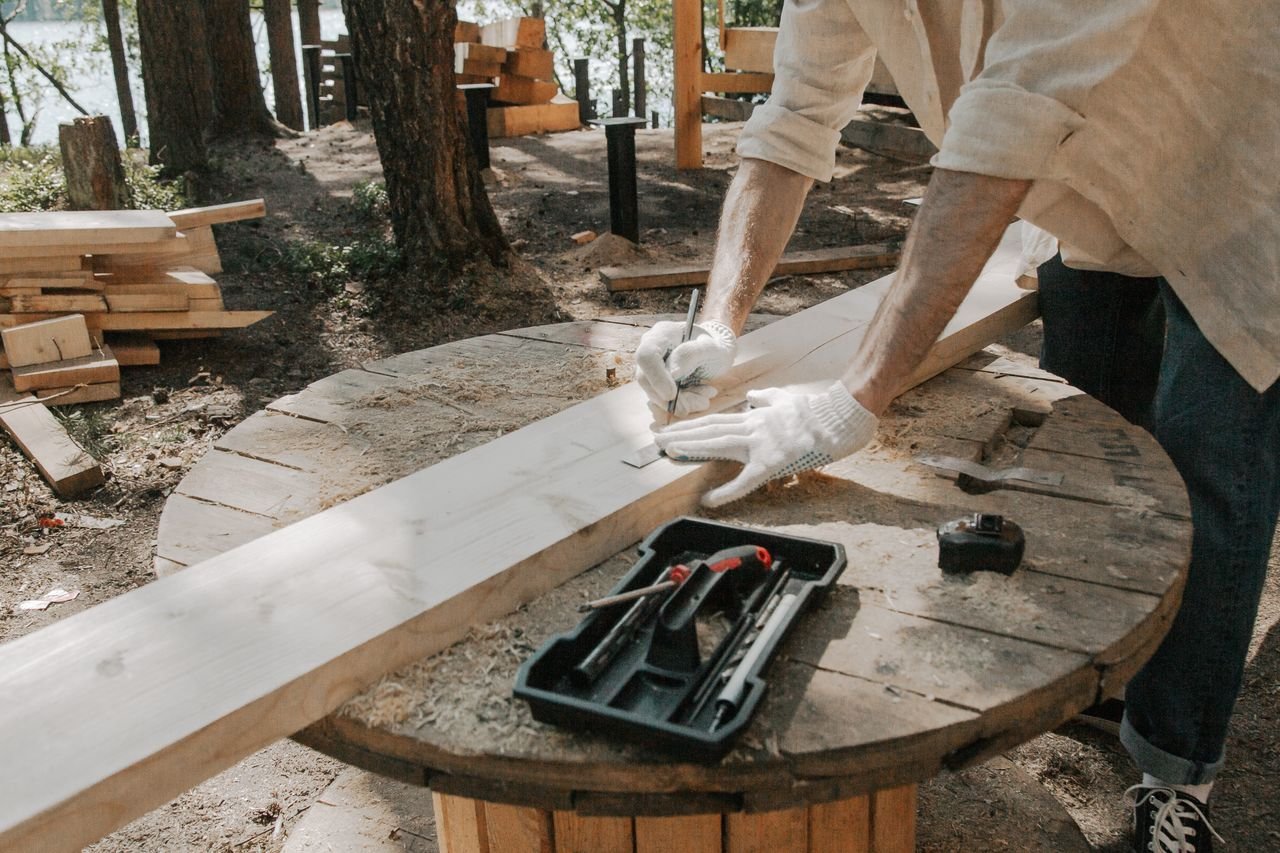 A carpenter skillfully outlines on a wooden plank with tools outdoors, surrounded by trees.